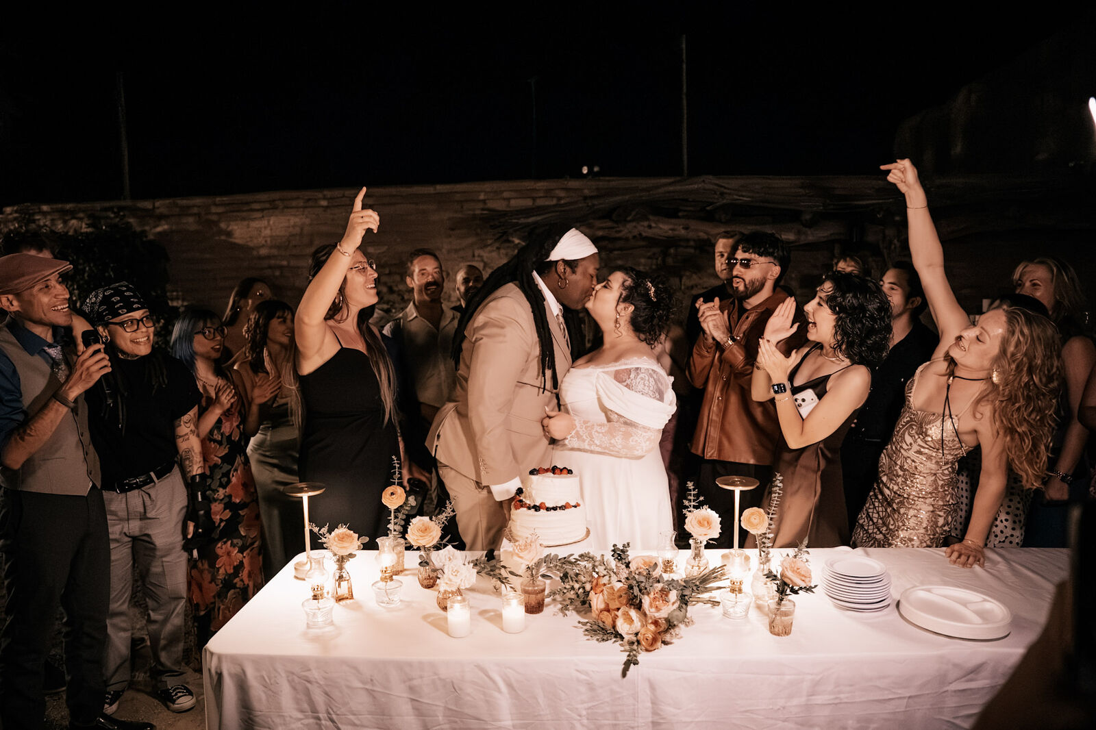 Guests cheering around the candlelit cake table at a night reception in the courtyard