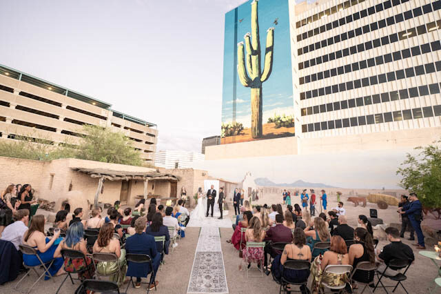 A packed golden-hour courtyard ceremony beneath the saguaro mural and the downtown skyline