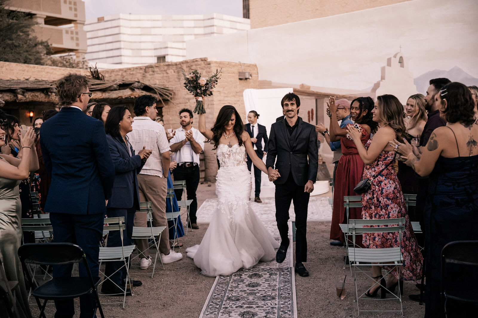 A just-married couple walking back up the aisle through a cheering crowd in the Presidio courtyard, downtown towers above the adobe