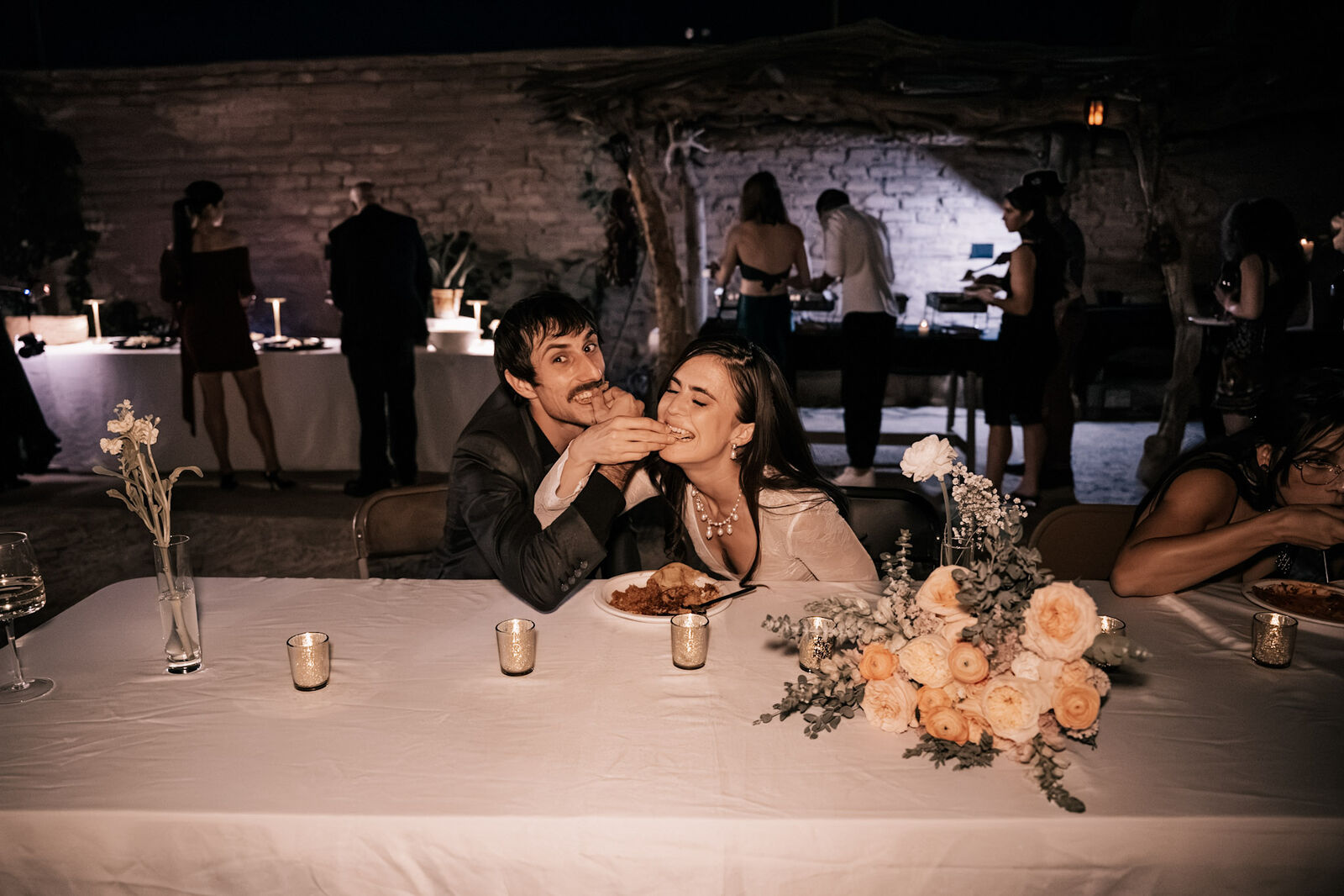 A newlywed couple feeding each other at a candlelit sweetheart table in the courtyard