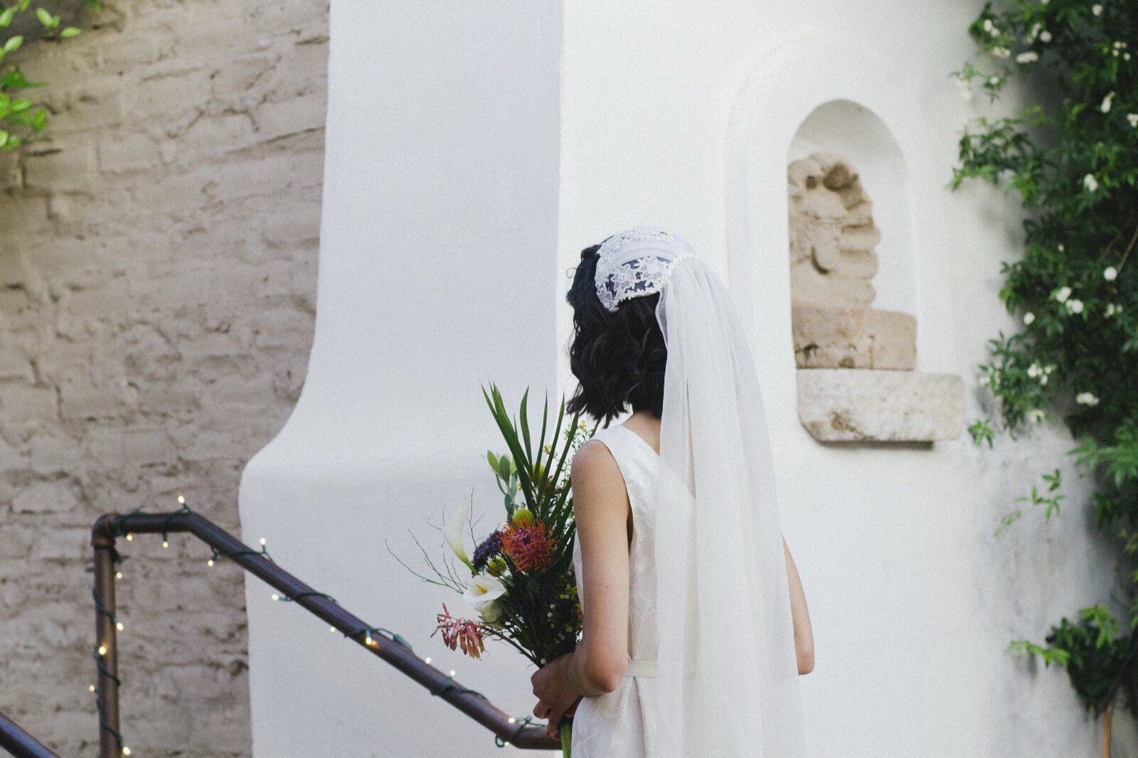 A bride in a lace veil beside the white plaster niche and climbing roses