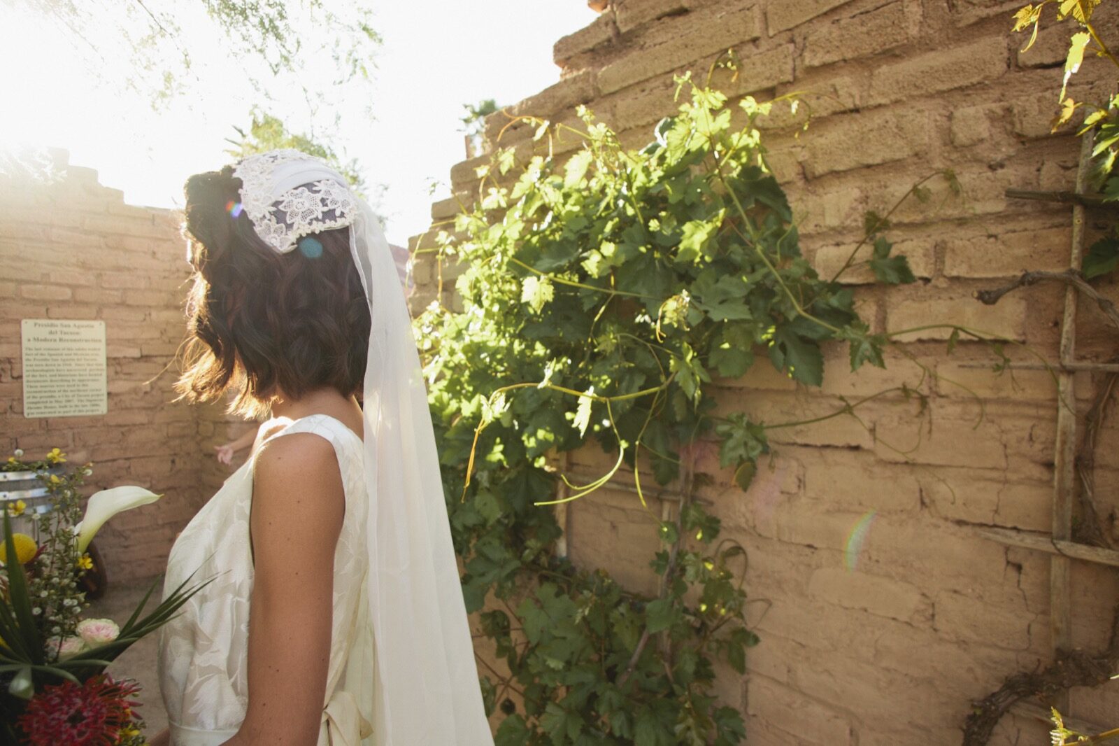 A bride in a long veil framed by the adobe wall and greenery