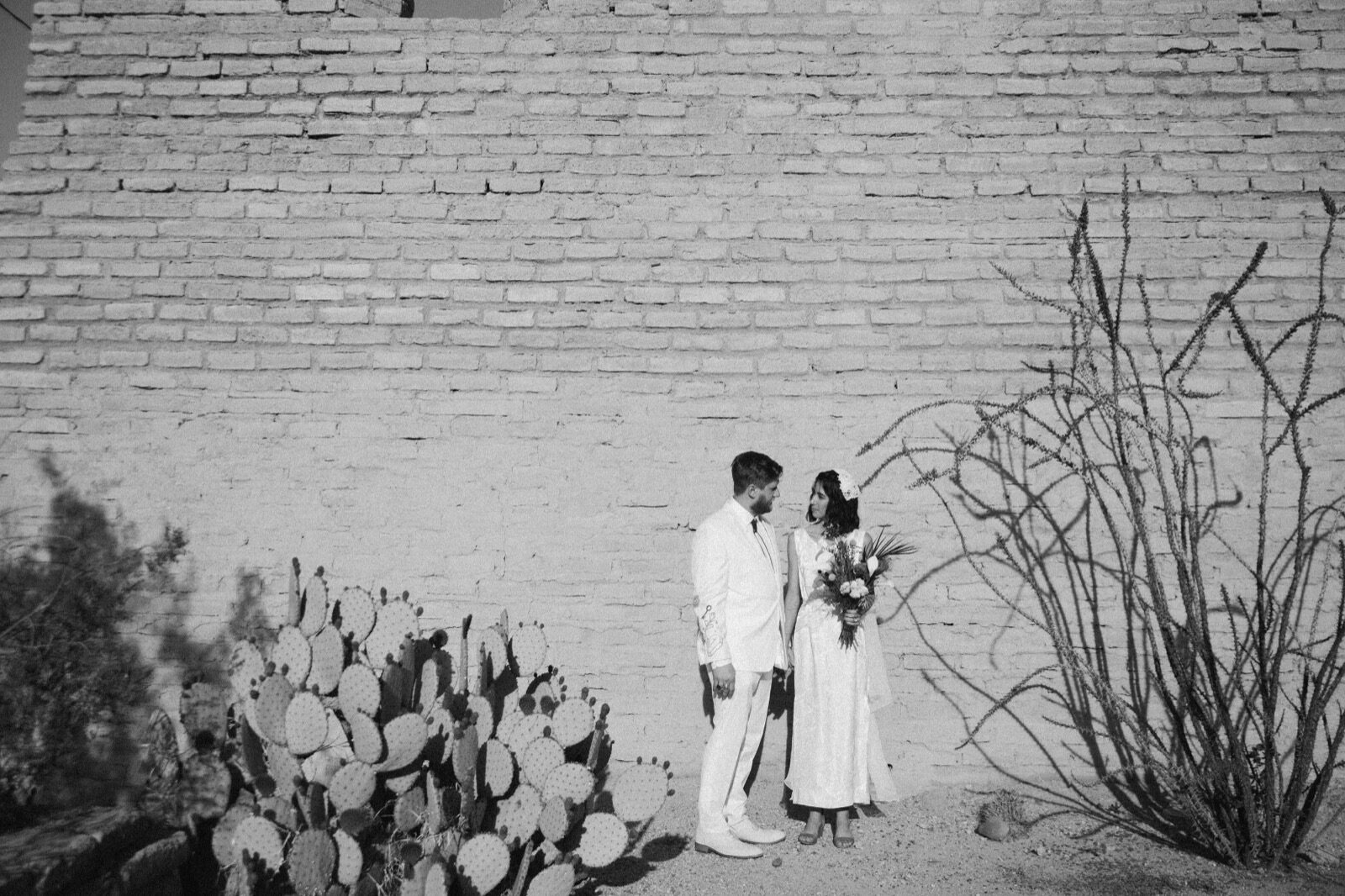 A black-and-white portrait of a couple against the adobe wall and desert plantings