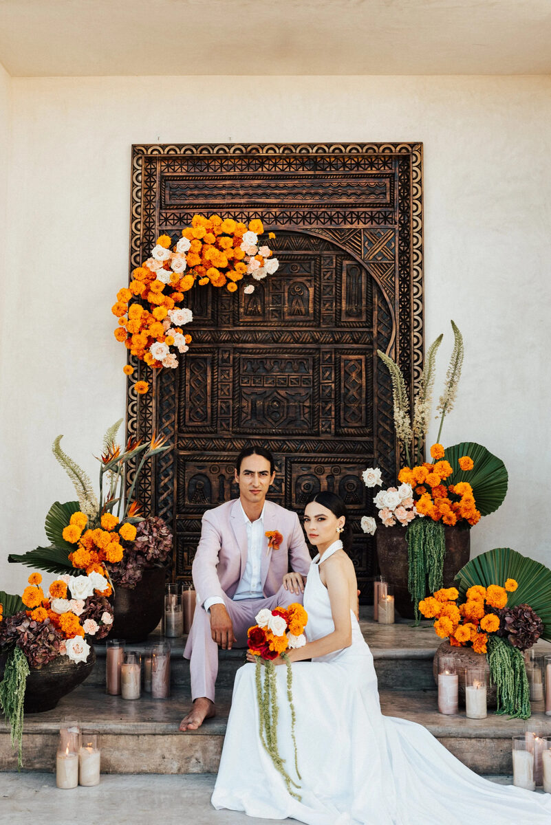 A couple seated before a carved wooden door framed in orange and marigold florals by Bloom Maven