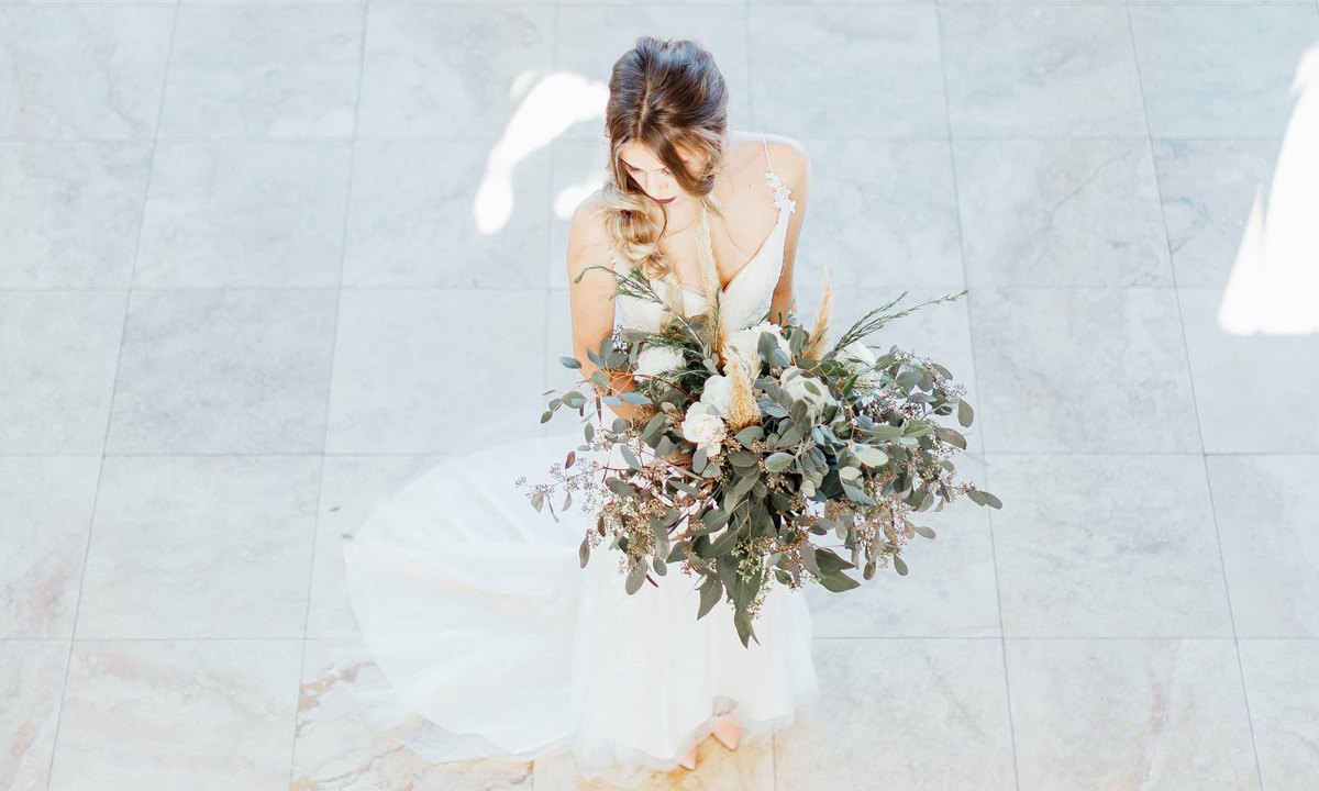 A bride in a flowing white gown holding a eucalyptus bouquet on a marble floor